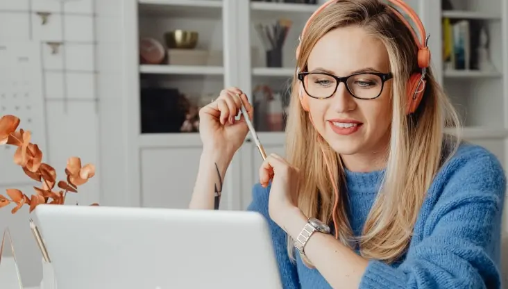 Woman with glasses and headphones smiling while looking at laptop with pen in hand