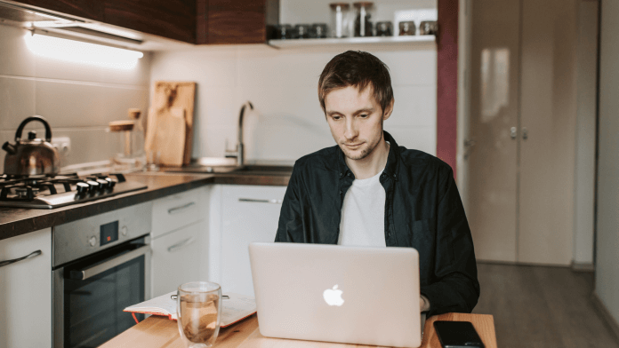 Young man using laptop at desk located in front of small kitchen area