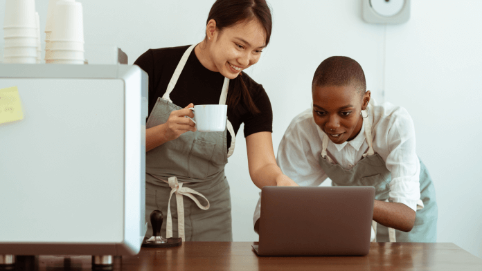 A woman in an apron pointing to something on the laptop that a kid is using.