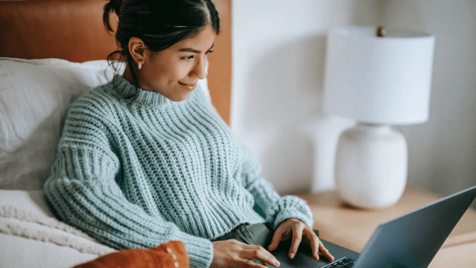 Woman wearing blue top is smiling while working with her laptop at the bed.