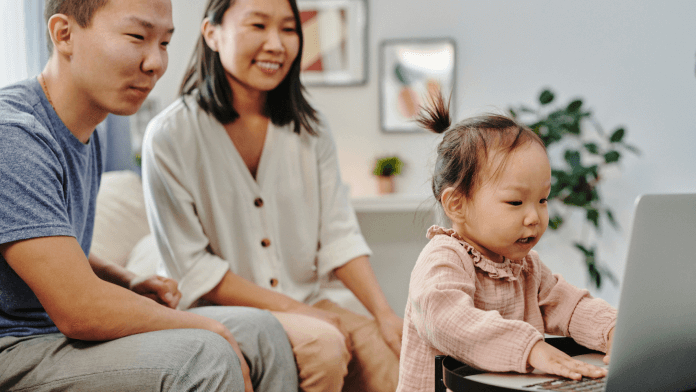 The toddler and parents are looking at a laptop, where parents are sitting on the couch.